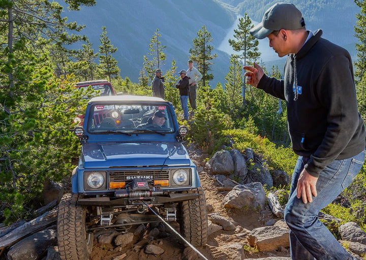 WARN vehicle recovery communication scene showing spotter guiding driver during winch operation with hand signals for safe off-road recovery